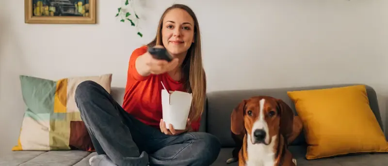 A woman sits on a sofa with her dog, holding a takeout box and pointing a TV remote.